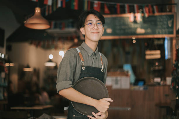 asian chinese teenage boy waiter looking at camera smiling work at cafe opening rear view asian chinese teenage boy waiter tying up apron getting read to work at cafe opening first job photos stock pictures, royalty-free photos & images