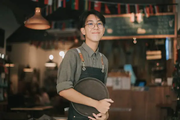 Photo of asian chinese teenage boy waiter looking at camera smiling work at cafe opening