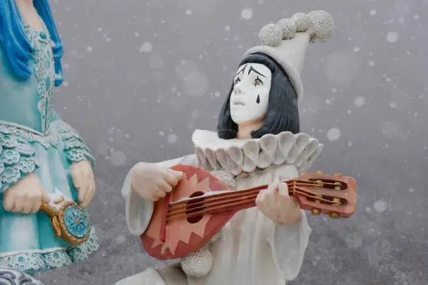 Statuette of boy in the image of Pierrot playing musical instrument in front of a girl during a snowfall. Unrequited love, problem relations concept.