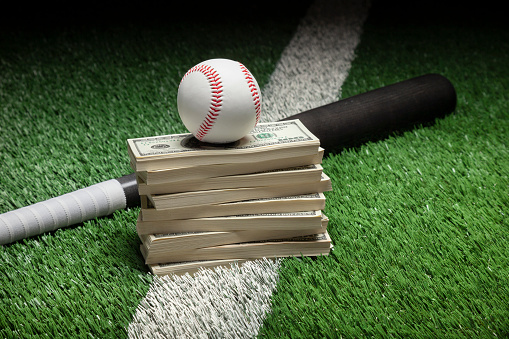 Baseball on top of stack of money with bat on green field with stripe
