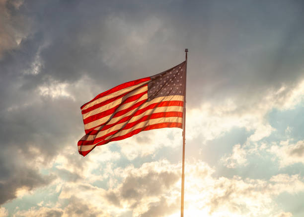istock Back lit American flag waving under clouds with sun rays at sunset 1291976979