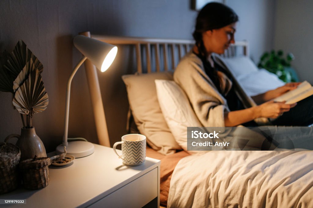 Hot beverage on a night stand Hot tea in a cup on a night stand. On the bed a woman is reading a book in cozy clothes. Night time, horizontal photo. Night Table Stock Photo