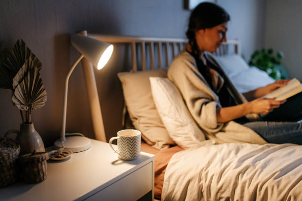 Hot beverage on a night stand Hot tea in a cup on a night stand. On the bed a woman is reading a book in cozy clothes. Night time, horizontal photo. night table stock pictures, royalty-free photos & images