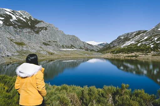 Mountain landscape with snowy mountains and lake. Woman with yellow coat looking at the lake. Lake Isoba, Leon. Spain.