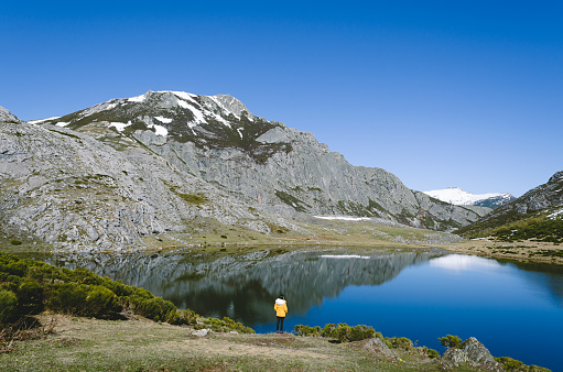 Mountain landscape with snowy mountains and lake. Woman with yellow coat looking at the lake. Lake Isoba, Leon. Spain.