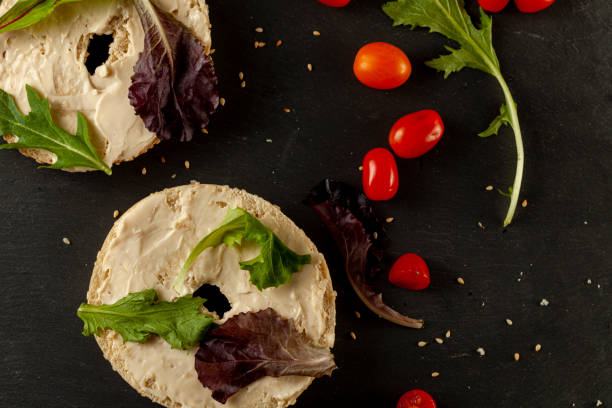 Toasted sesame bagel buttered with cream cheese served with arugula and cherry tomatoes Toasted and sliced sesame bagel with creamy spread on it served with fresh greens (arugula) and cherry tomatoes on black wooden background. A healthy snack alternative or breakfast idea. heathy snack stock pictures, royalty-free photos & images