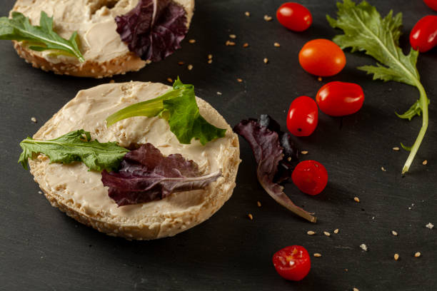 Toasted sesame bagel buttered with cream cheese served with arugula and cherry tomatoes Toasted and sliced sesame bagel with creamy spread on it served with fresh greens (arugula) and cherry tomatoes on black wooden background. A healthy snack alternative or breakfast idea. heathy snack stock pictures, royalty-free photos & images
