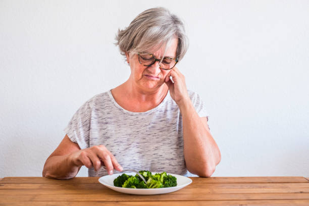 一位老人厭惡地看著家裡木桌上盤子里的花椰菜 — — 試圖節食和減肥 - 厭惡 個照片及圖片檔