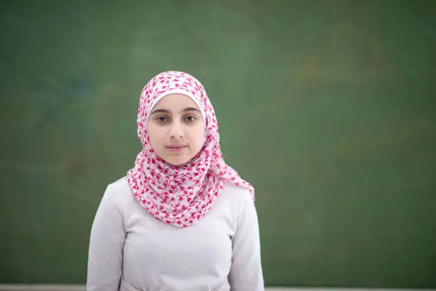 A serious looking Muslim elementary school student looks at the camera with a neutral expression. She is wearing a pink hijab.