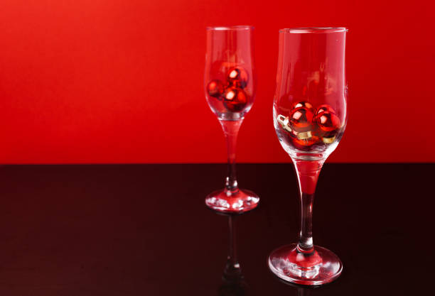 Two champagne glasses with small Christmas balls inside against red background close up
