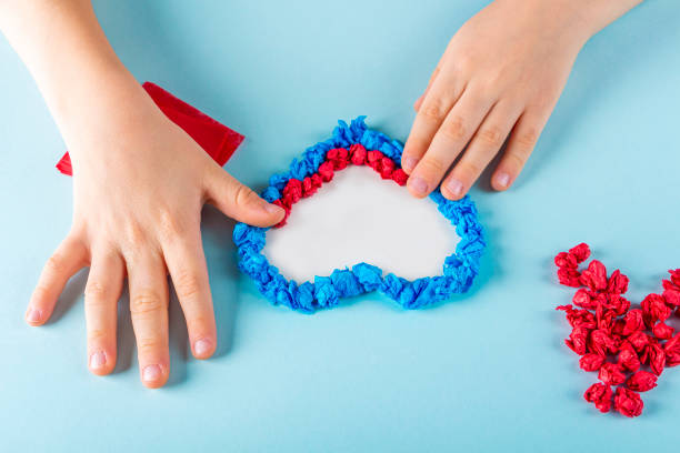 Concept of DIY and kid's creativity. Step by step instruction: make  heart. Step3 Child's hands stick crumpled pieces of paper on adhesive heart template. Valentine's Day craft. Fine motor skills