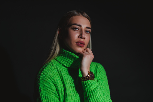 Portrait of a beautiful young fashionable woman with blond hair wearing a green sweater against a black background