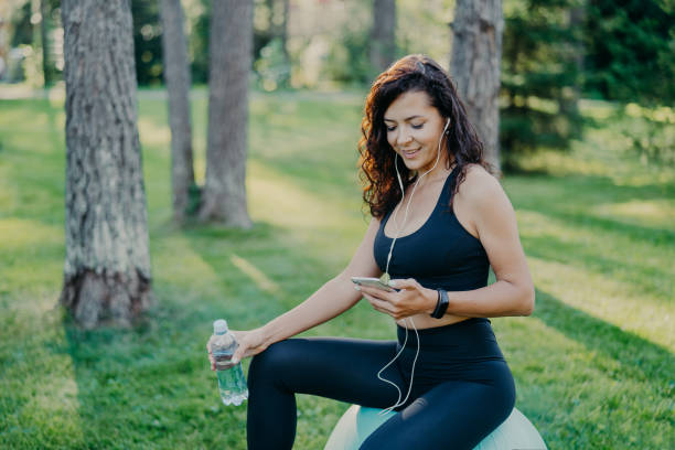 Pleased relaxed woman has rest after yoga exercises sits on fitness ball, listens music via modern mobile phone and earphones, holds bottle of fresh water, dressed in active wear, poses at park mujer relajada y complacida tiene descanso después de ejercicios de yoga se sienta en la pelota de fitness, escucha música a través de teléfono móvil moderno y auriculares, sostiene botella de agua dulce, vestido con ropa activa, posa en el parque