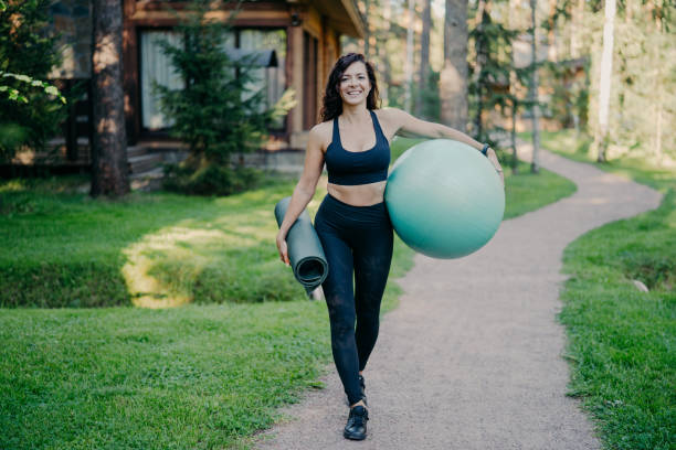 Outdoor shot of happy sporty brunette woman walks on path near forest, holds rolled up karemat and fitness ball, dressed in cropped top, leggings and sneakers, has regular trainings in open air tiro al aire libre de mujer morena deportiva feliz camina en el camino cerca del bosque, sostiene karemat enrollado y pelota de fitness, vestido con la parte superior recortada, leggings y zapatillas, tiene entrenamientos regulares al aire libre