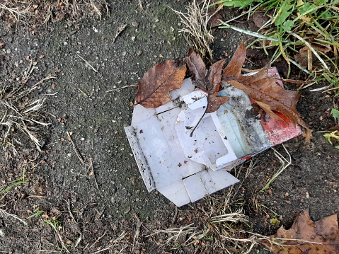 An empty cigarette pack or box trown away outside. Wet paper and plastic, some keaves, grass anf dirt.
