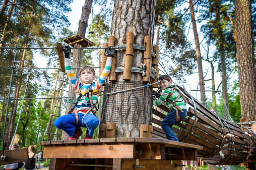 Two little boy boys kid in a helmet and with a safety carbine goes on a rope on the background of forest and sky. Healthy games outdoor.