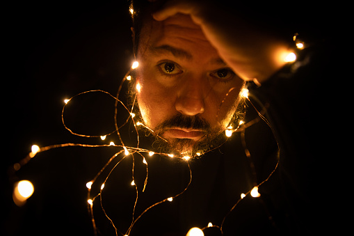 Young man holds his head with a worried face entangled by a garland of lights. Sad moments concept