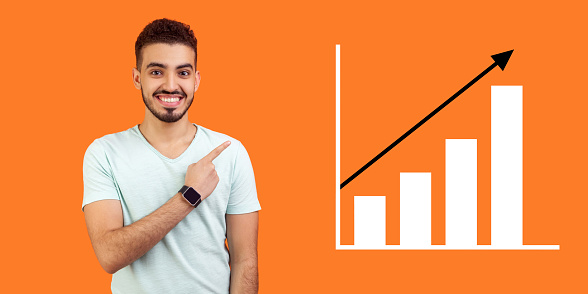 Portrait of happy joyful young bearded man standing, pointing aside and showing business growth graph. indoor studio shot isolated on orange background