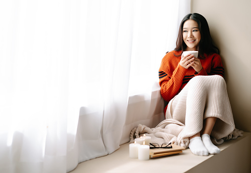 Young beautiful happy Asian woman wearing knitted warm sweater sitting home in the chair by the window with cup of hot drink at home.Cozy winter decorated with candles.Feeling comfortable at home.