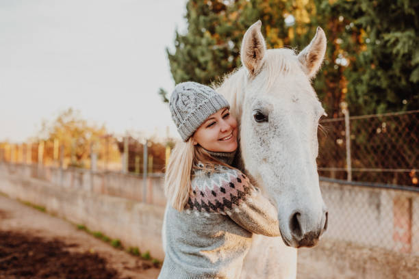 istock Blond young girl with a woollen cap and old winter pullover embracing her white mare in the horse stable 1292133062