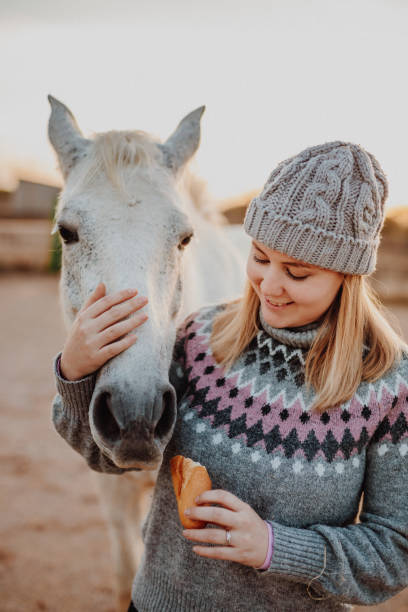 istock Smiling young blond woman with a woollen cap and old winter pullover feeding her white mare with dry bread in the horse stable 1292133498