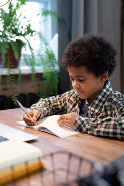 istock Cute little African schoolboy with pen making notes on page of copybook 1292135630