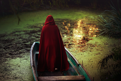 Rear view of woman in red cape standing in wooden boat looking away at the lake