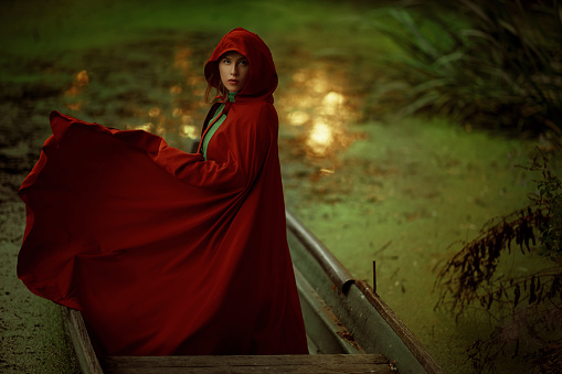 Woman wearing flying red cape posing at the green lake looking at camera