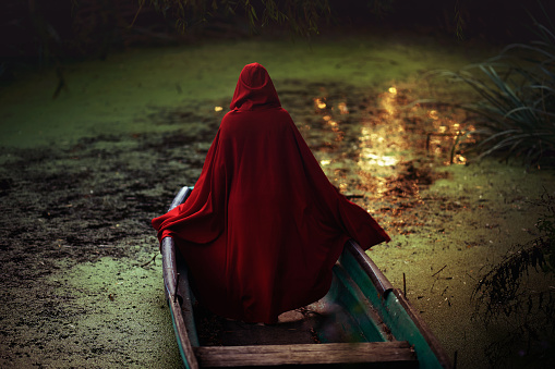 Woman standing wearing red cape at the lake in wooden boat, feeling mysterious