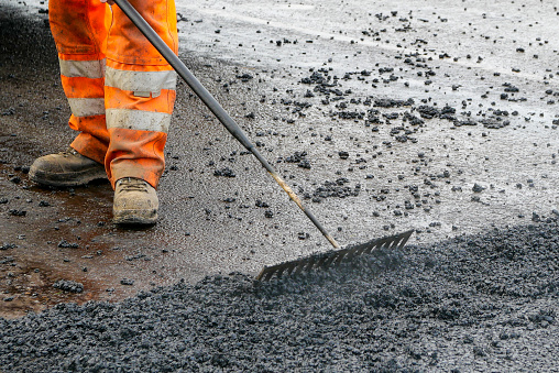 A construction worker in bright orange reflective workwear raking loose  asphalt stones into position. He is moving the sticky black chippings into an area of new road surface, before it is rolled and levelled. He is wearing heavy duty work boots to protect his feet on a busy construction site.