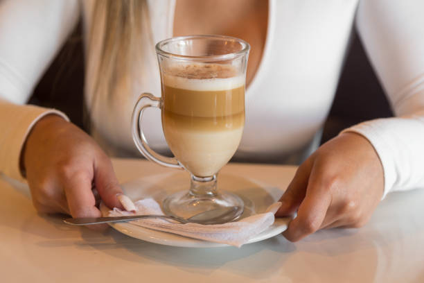 copo de vidro com café e creme, em um prato com uma colher em uma mesa de madeira, realizada por duas mãos - cream liqueur - fotografias e filmes do acervo