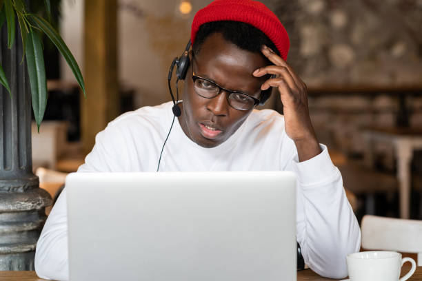 thoughtful millennial black man in cuffia indossa il cappello rosso, guardando webinar educativi su laptop, lavoro online remoto nel caffè. audio lezione o lezione di ascolto maschile afro-americano. e-learning. - the thinker audio foto e immagini stock