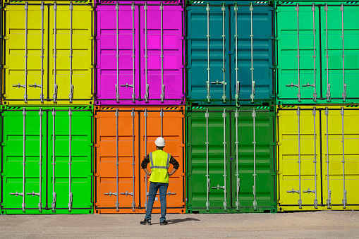 Engineer man with yellow crash helmet and worker west checking cargo freights in front of colorful cargo container stacks in shipping port.