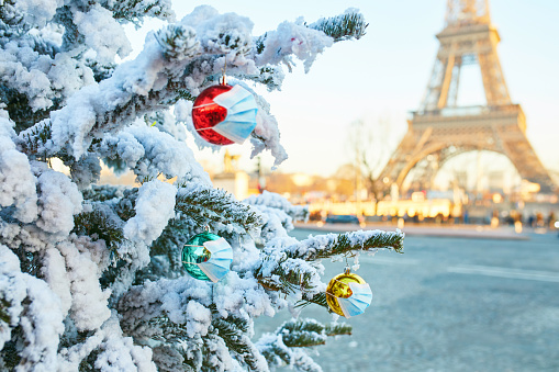 Christmas tree covered with snow and decorated with red, green and yellow balls wearing face masks, Eiffel tower in the background. Season holidays during pandemic and lockdown in Paris, France