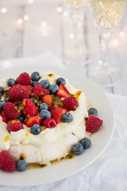 Pavlova with strawberries, blueberries, raspberries and passionfruit with Christmas lights and sparkling wine in the background to celebrate the holidays in Australia