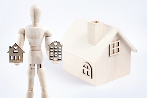 A wooden figure of a symbolic person holds a small house in his hands against the background of a toy house