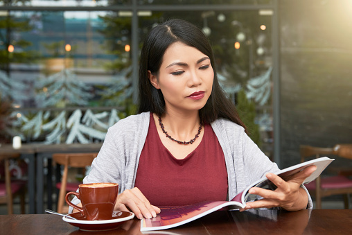 Pensive young pretty Asian woman drinking cup of coffee and reading magazine in coffeeshop