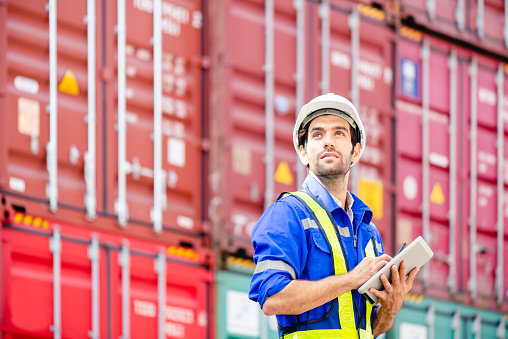 Engineer or Technician wears white hardhat and reflection shirt and holding tablet and look at sky and smile with cargo stacked containers in background. Concept of inventory and logistic management.