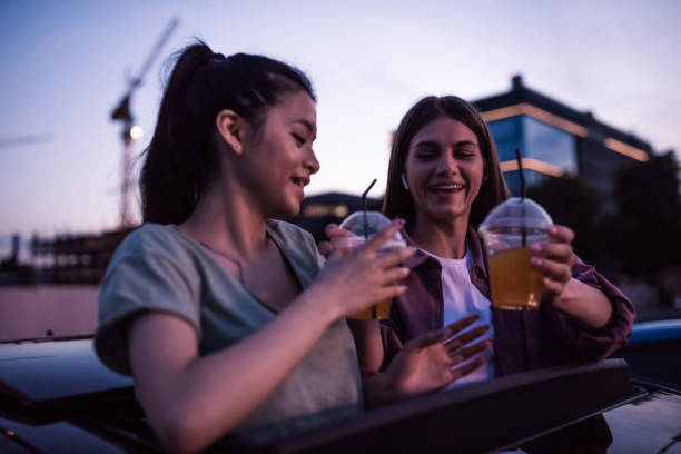 duas garotas parecendo alegres, segurando bebidas enquanto sentam no carro e assistem a um filme em um cinema ao ar livre à noite - outdoors drinking women friendship - fotografias e filmes do acervo