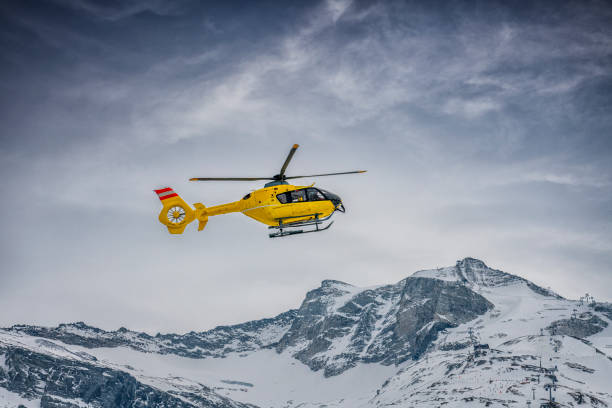 A red rescue helicopter in flight against a deep blue sky, soaring above a grassy mountain slope. The dramatic angle emphasizes speed and urgency, symbolizing emergency response and aerial operations.