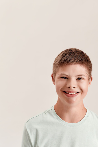 Close up portrait of cheerful disabled boy with Down syndrome smiling at camera while posing isolated over white background. Children with disabilities and special needs concept. Vertical shot