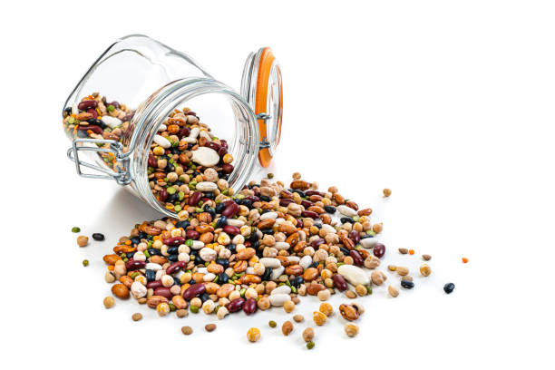 Food Backgrounds: large variety of multicolored dried mixed beans and legumes spilling out a glass jar isolated on white background. The composition includes green, yellow and brown lentils, chick-peas, black beans, Pinto beans, Kidney beans, fava beans, white beans, mung beans and soy beans among others. High resolution 42Mp studio digital capture taken with SONY A7rII and Zeiss Batis 40mm F2.0 CF lens