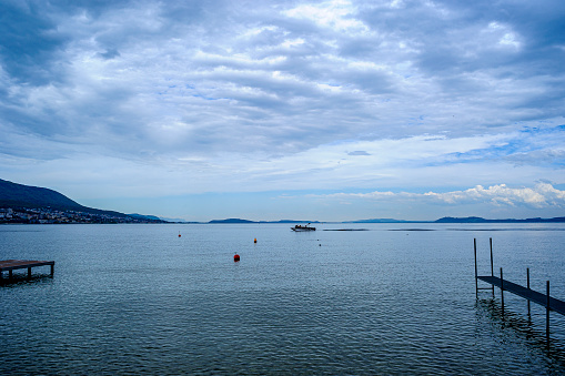 Photo taken with a wide-angle lens. View of Lake Neuchâtel at blue time. The perspective makes two footbridges converge towards a boat passing in the background.  Cloudy sky over the lake.