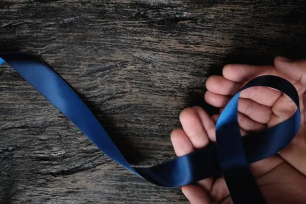 Photo of Top view of male hand holding dark blue ribbon on wooden background with copy space. Colon, stomach, colorectal cancer, arthritis, ards acute respiratory distress syndrome awareness and child abuse prevention concept.