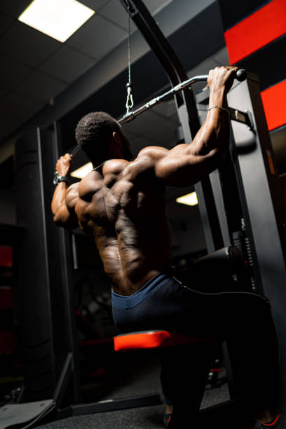 istock Athlete pumps biceps on the simulator. African handsome bodybuilder in gym. View from the back. 1292228671