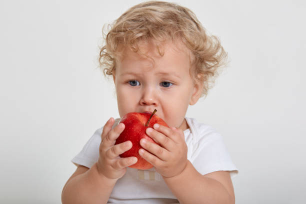 istock Baby with apple posing isolated over white background, blond tot biting red fruit and looking away, vitamins for male kid, wavy haired child having snack. 1292234513