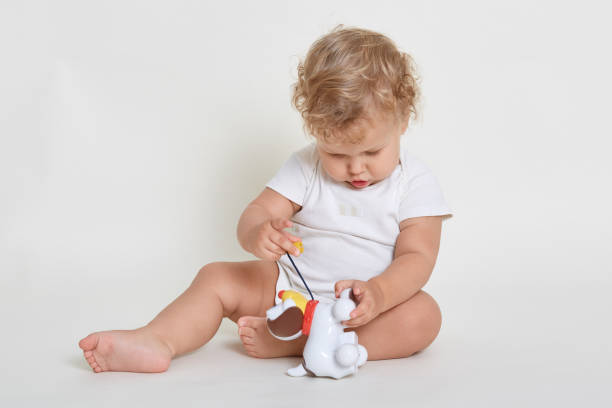 istock Baby boy dresses white bodysuit posing isolated over light background, sitting on floor, playing with plastic dog, child with blond wavy hair plays indoor. 1292234526