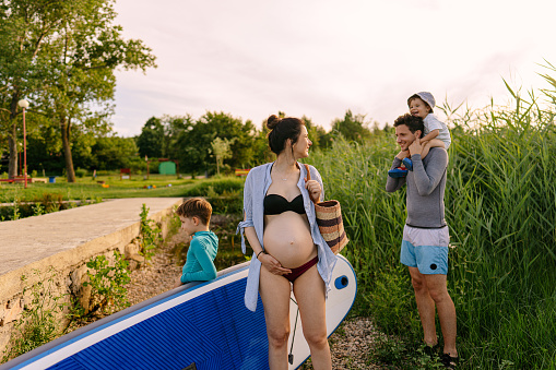 Photo of a young family on a summer vacation: two little boys and father are about to ride a paddle board, while mother is pregnant and expecting another child