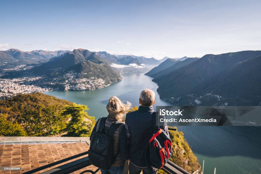 Mature couple hike above lake Lugano in the morning They relax at viewpoint and look off to distant scene Travel Stock Photo