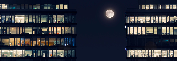 Two office buildings with many lit windows with a full moon on the night sky between them.